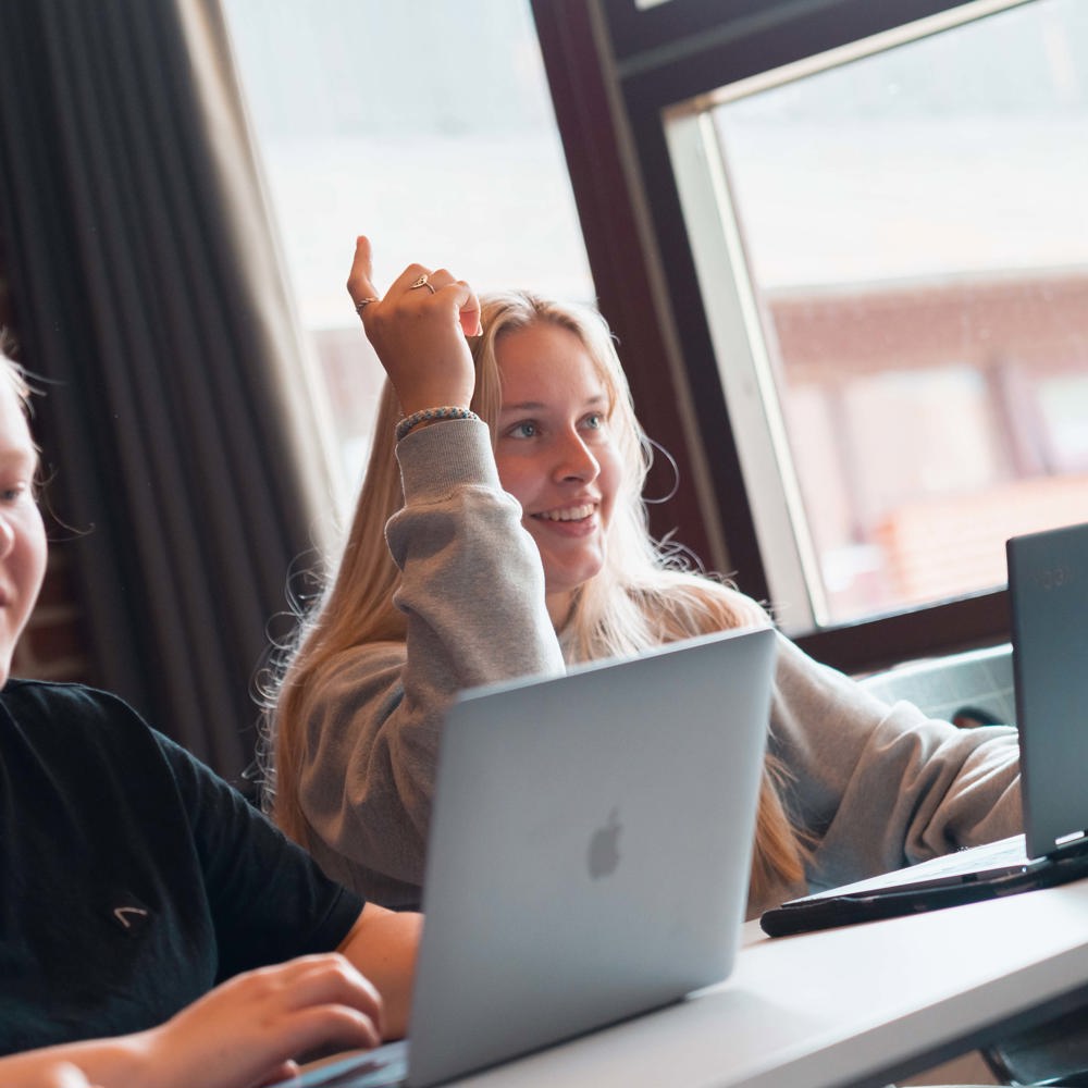 To elever rækker hånden op i et klasseværelse, mens de arbejder ved laptops