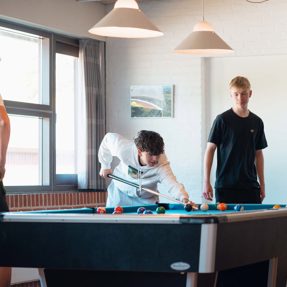 Tre unge spiller pool i et opholdsrum med store vinduer og lyse lamper